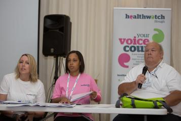 Man and two women sitting at a desk, the man is talking into a microphone