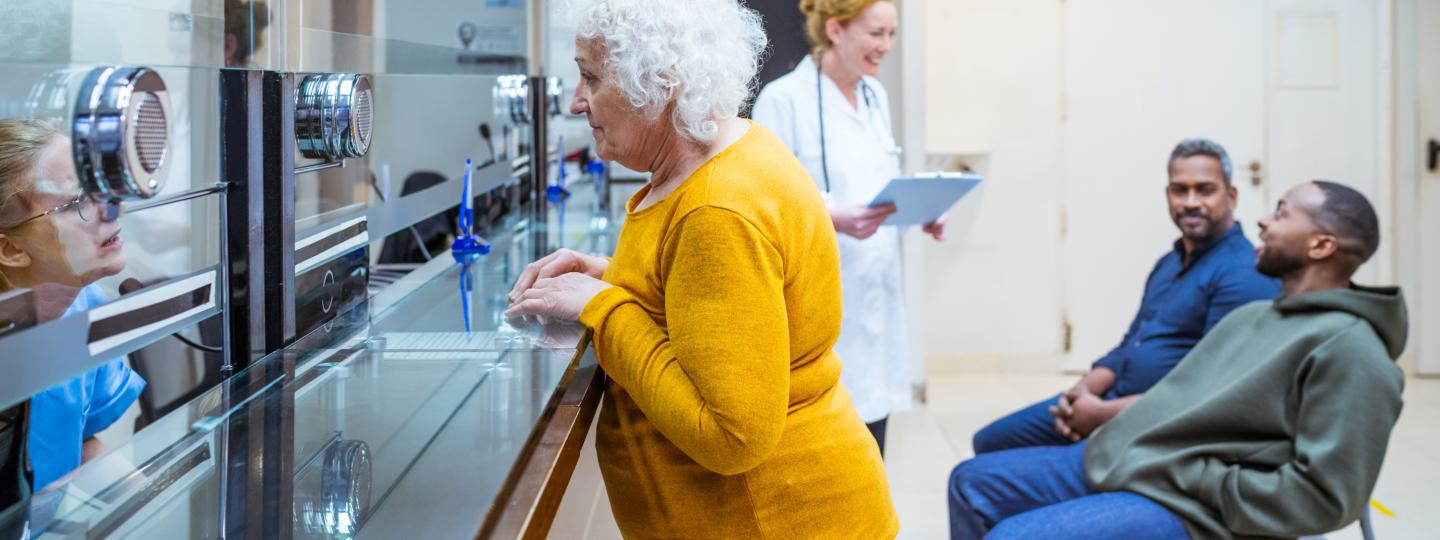 Senior woman talking with receptionist in waiting room in medical clinic. Two men talking with female doctor in the background.