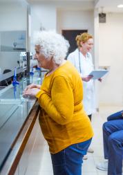 Senior woman talking with receptionist in waiting room in medical clinic. Two men talking with female doctor in the background.