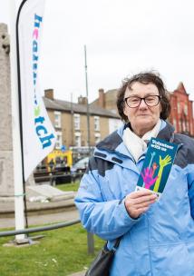 Woman stood in front of a Healthwatch flag holding up a leaflet about volunteering